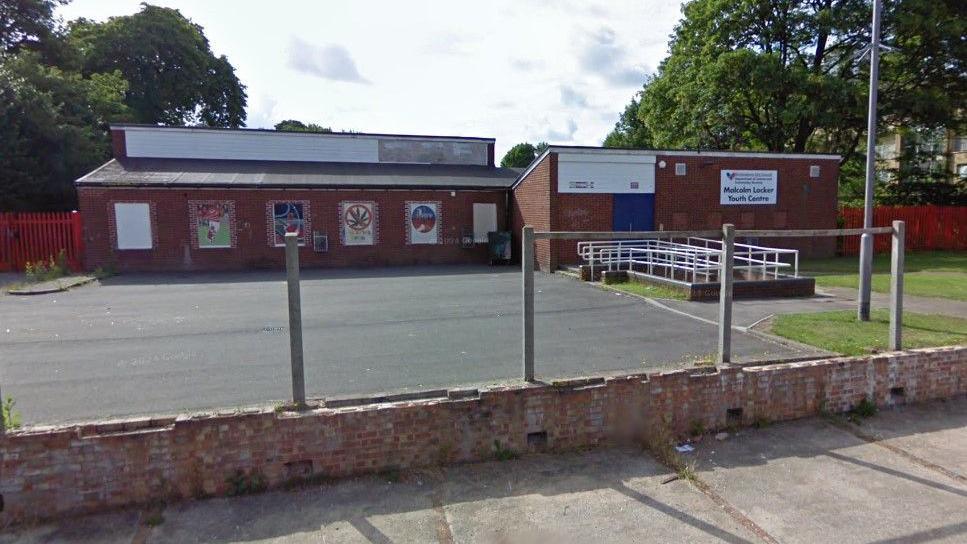 A single-storey brick building with a tarmac yard at the front. There is artwork on the walls and a Birmingham City Council sign reading "Malcolm Locker Youth Centre" 