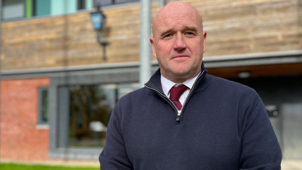 A bald man looking at the camera. Behind him is a building. He is wearing a blue fleece with a zip open. Under that he has a white shirt and burgundy tie.