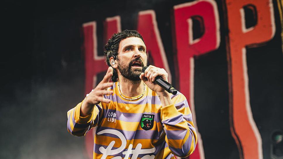Sergio Pizzorno of Kasabian performs onstage during Tramlines at Hillsborough Park. He wears a yellow and purple striped jersey.