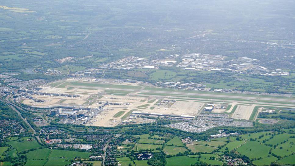 An areal image of Gatwick Airport. There is green fields on either side of the large airport. 