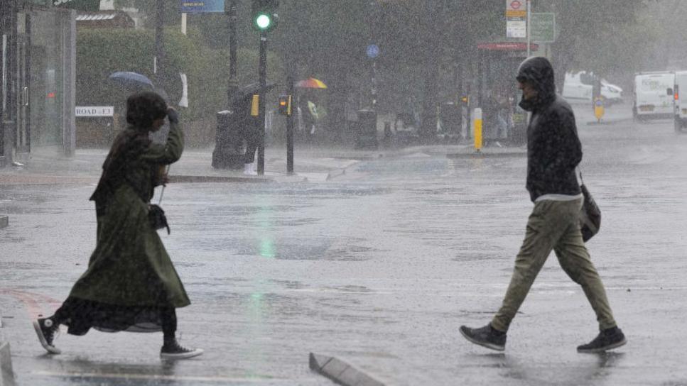 Two people crossing an urban road in heavy rain, one shielding their face from the rain