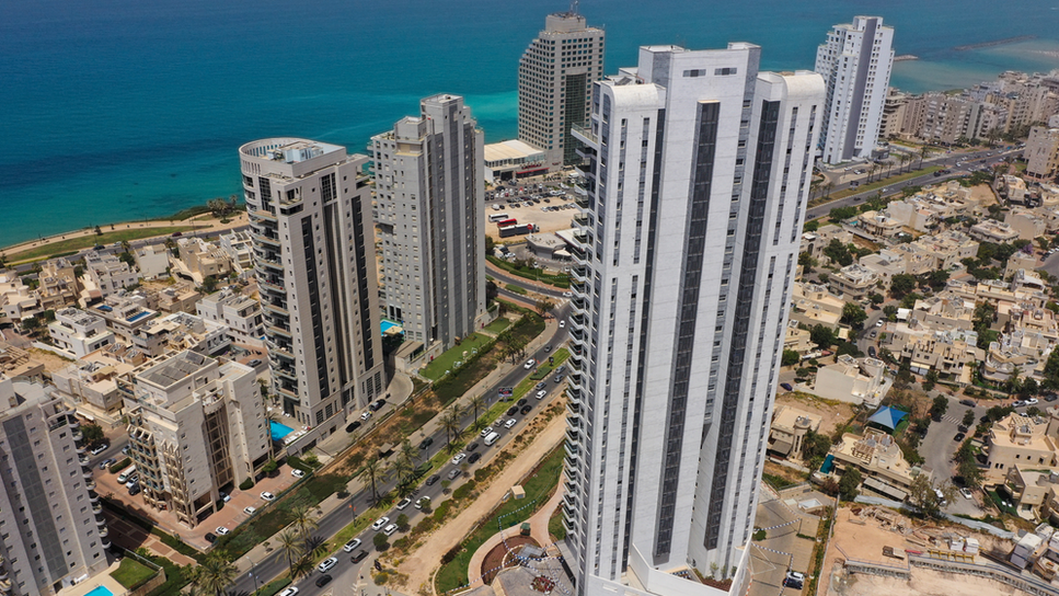 Aerial photo of Netanya, a built-up area with several skyscrapers. Palm trees can be seen on the road. The city is next to the sea, which is very blue.