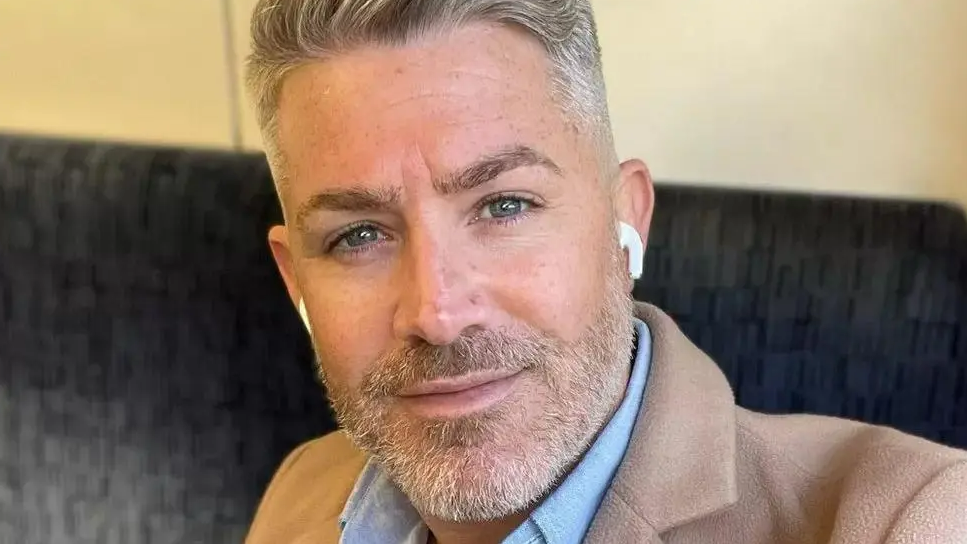 Michael Porter looking directly at the camera. He has grey hair which is up in a quiff at the front. He has dark eybrows and blue eyes. He has short grey and white beard covering the lower part of his face and is wearing white wireless earbuds in his ears. He is wearing a blue shirt, the collar of which is touching his chin, under a beige jacket.