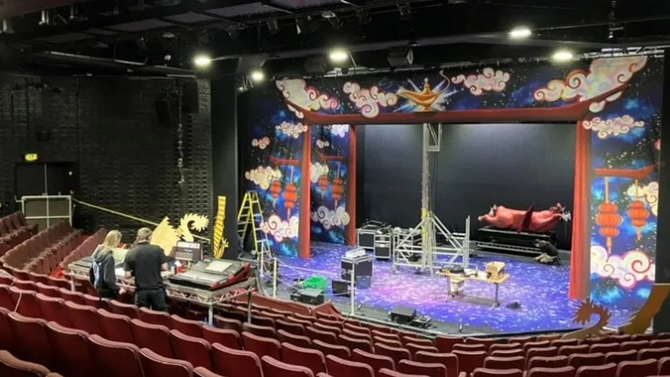 Inside the theatre with rows of red seats and a black stage with colourful props 