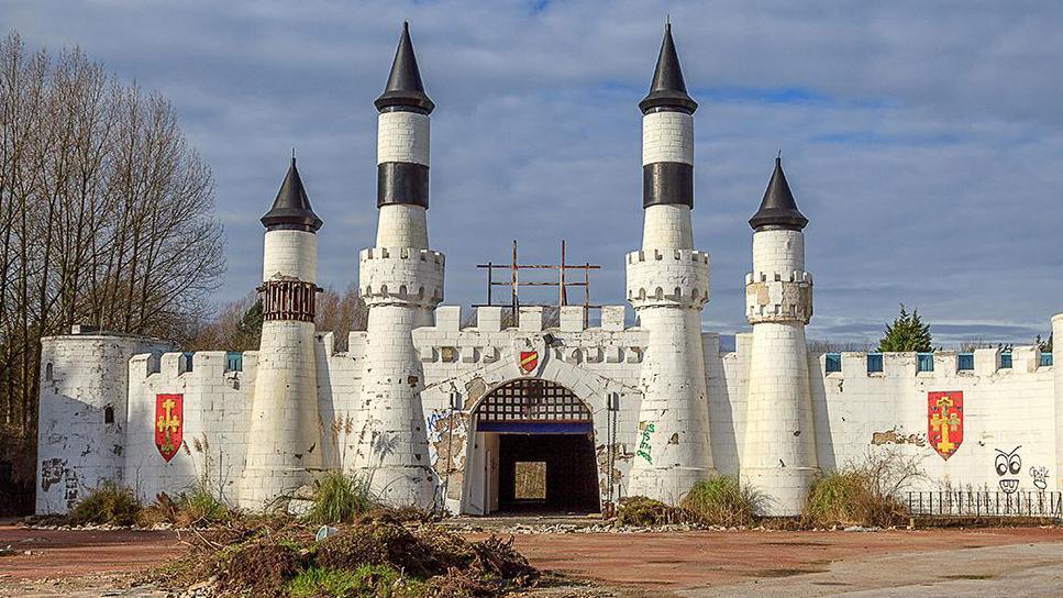 The entrance to the abandoned Camelot theme park. The image shows a mock white castle frontage with a barbican and turrets. There are coats of arms on the walls and overgrowth in the foreground.