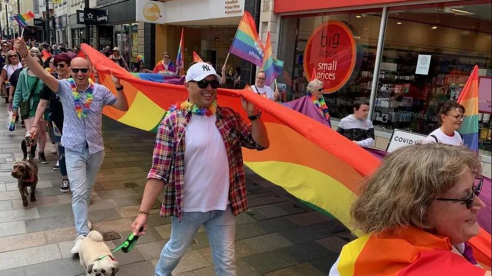 People walk down a street of shops holding up a giant long rainbow flag. They also wear colourful clothes and wave other smaller flags