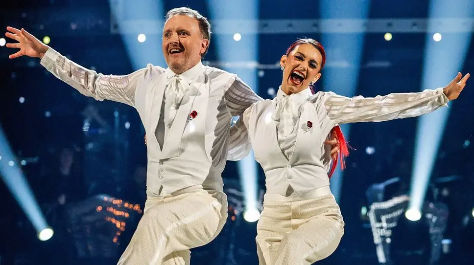 Comedian Chris McCausland and dancer Diane Buswell, a woman with long dyed red hair, perform a dance routine wearing white suits.