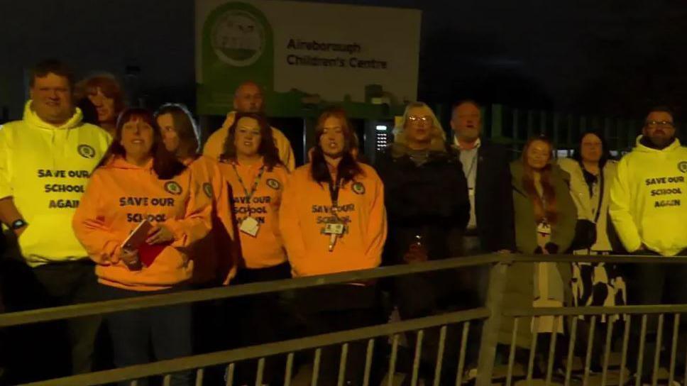 Several parents in orange and yellow hoodies line up outside the school in protest at its closure 