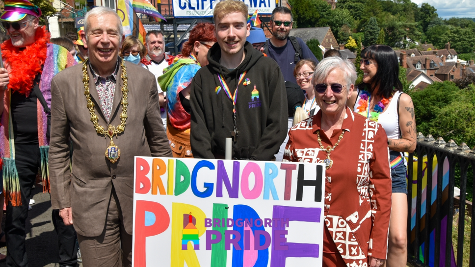 Huw has short blonde hair and is wearing a black hoodie. He jointly holds a Bridgnorth Pride poster, along with mayor David Cooper and mayoress Nicky Cooper.