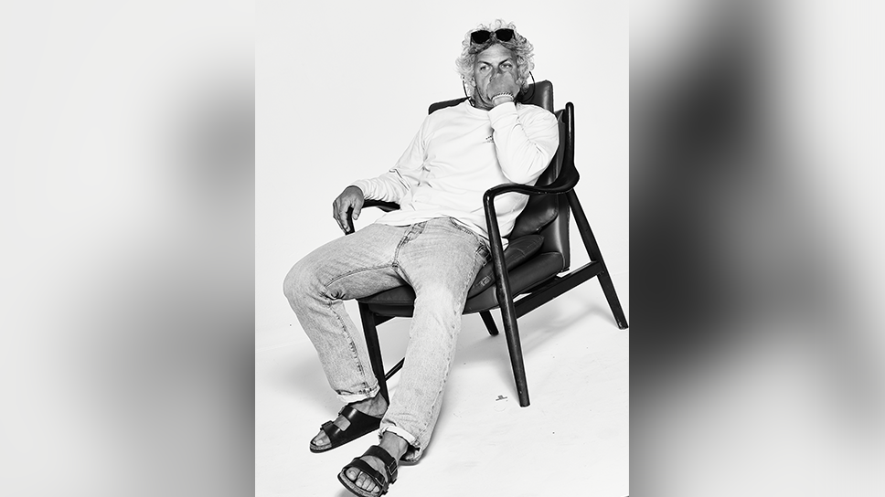 Tom Jackson in a black and white shot. He is lounging in a black leather chair in front of a white background. He's wearing jeans, no socks and Birkenstock sandals with a white cotton long sleeved round neck top. He has his hand up in a fist in front of the lower half of his face. He had long curly black and white hair and has a pair of dark sunglasses perched on his head.