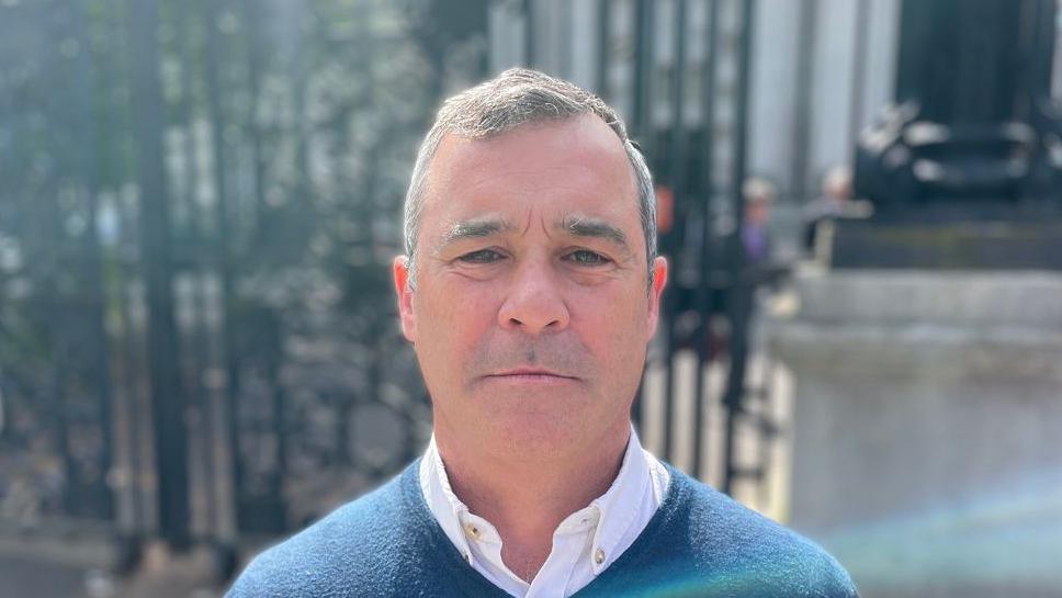 Alan Brecknell has short greying hair and is wearing a blue v-neck jumper with an open necked white shirt