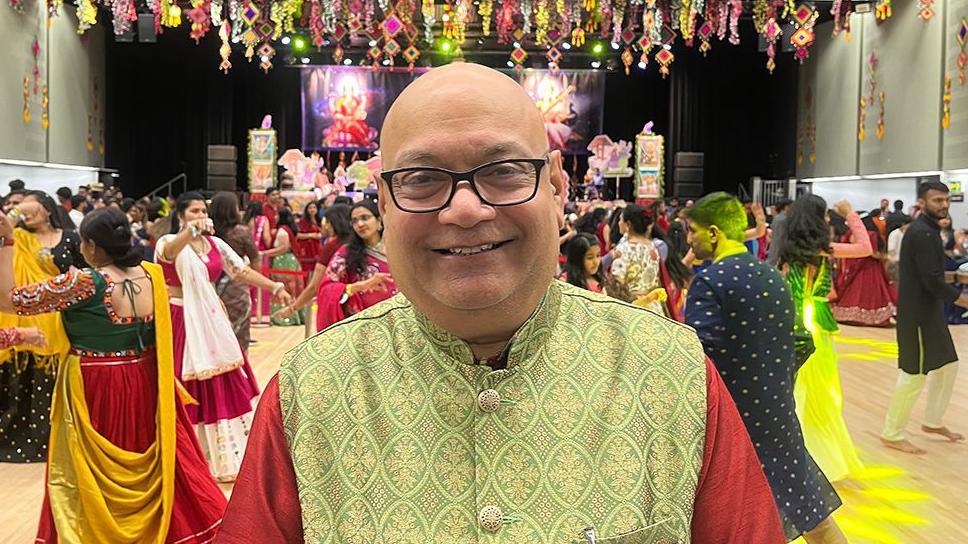 Festival organiser Sunil Chauhan wearing glasses and a green traditional Navratri outfit smiling as worshippers dance behind him