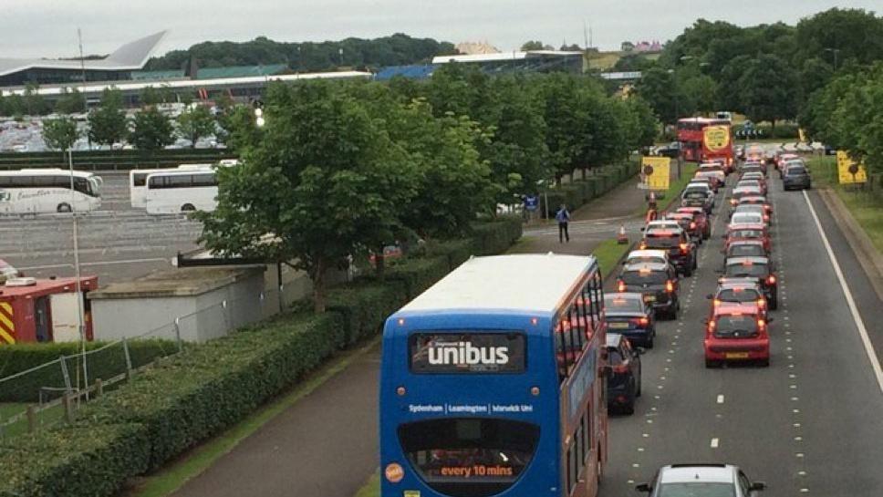 Approach road to Silverstone (Dadford Road). A queue of traffic including a blue bus as well as cars, is queuing towards a roundabout. There are temporary yellow signs near the roundabout. The carriageway is separated from the circuit by a hedge and a line of trees. White buses are visible in the car park to the left, along with some buildings.