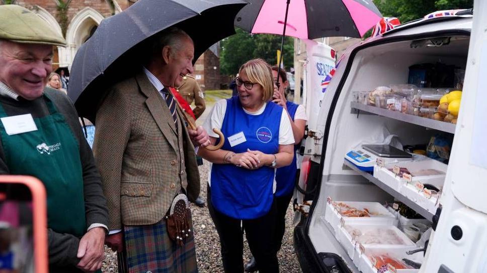The King wearing a kilt holding an umbrella, he is talking to a woman with blonde hair, glasses and a blue apron on. They are standing next to a white van which has seafood inside it.