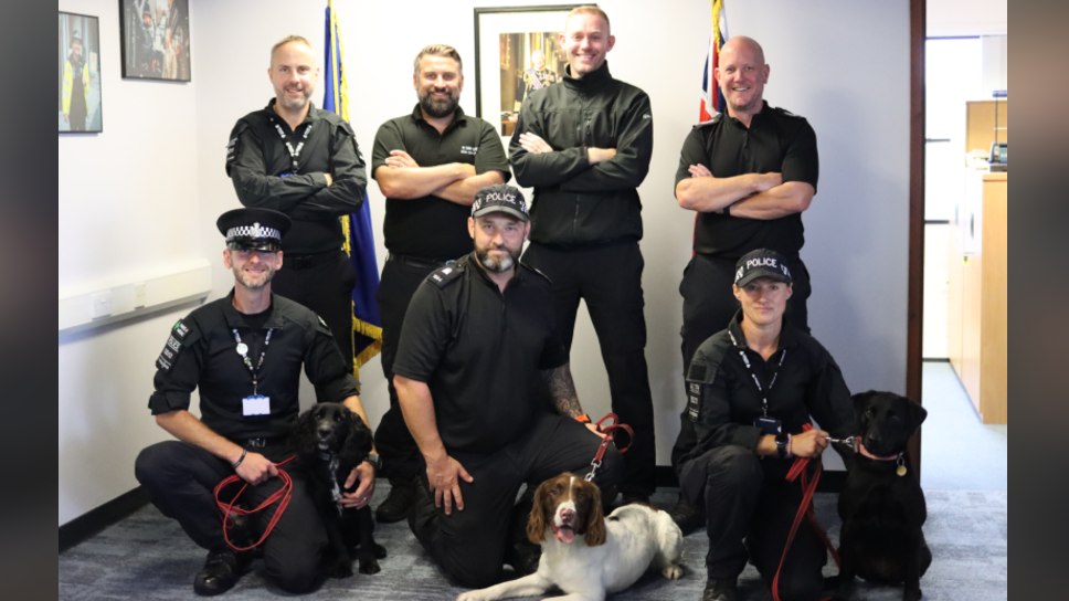 A group of seven police officers and three police dogs. On the back row is four officers and on the front row is three officers with the three dogs. All officers are looking at the camera and smiling and are all wearing police uniform. 