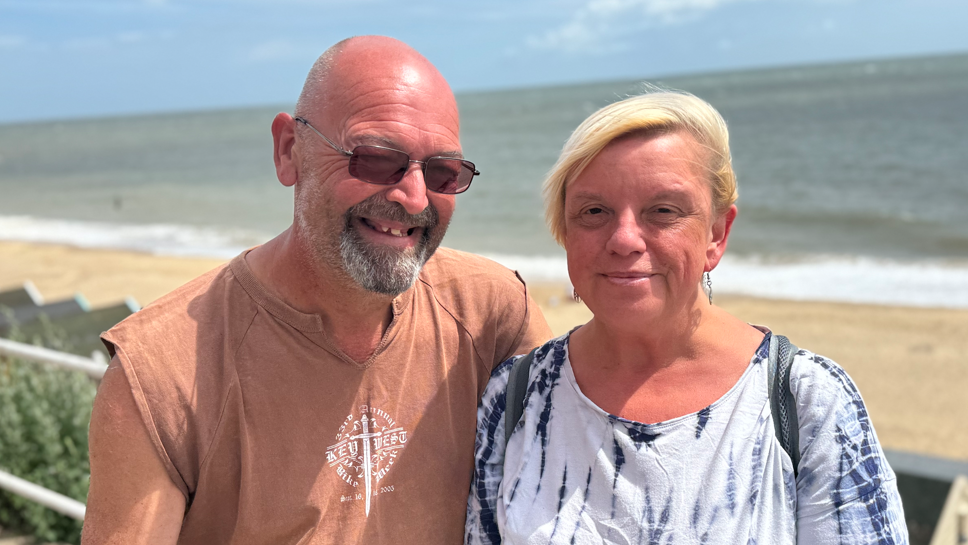 A head and shoulders image of married couple Nessa and Martin Howen. They are standing close together and looking directly into the camera. The beach and sea can be seen in the background.