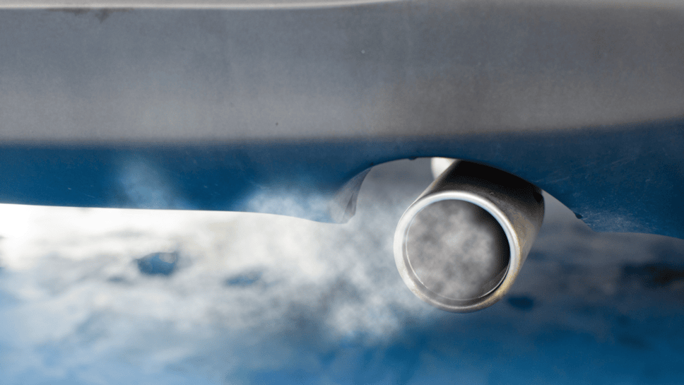 Stock image of a car exhaust. It is silver, at the back of a car and there is white smoke around the vehicle and a blue tint to the image.