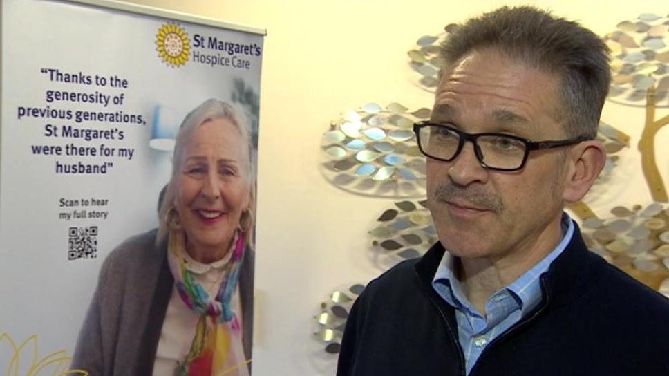 A man wearing glasses and a blue shirt looks at the camera, standing next to a St Margaret's Hospice banner 