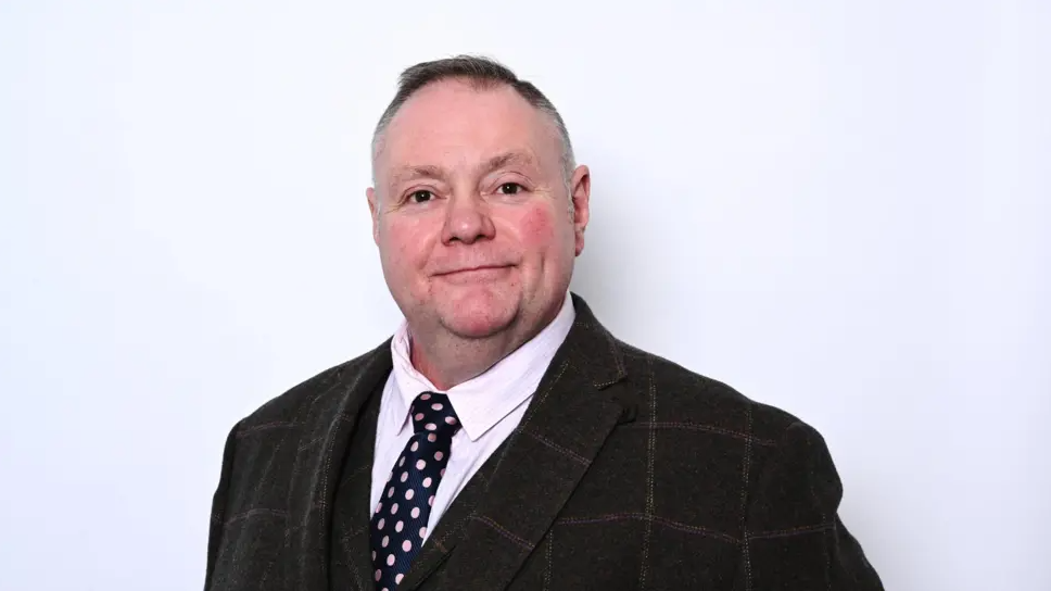 Mr Simkins is wearing a dark checked suit and pink shirt and dark tie. He is smiling at the camera and had short dark and grey hair.