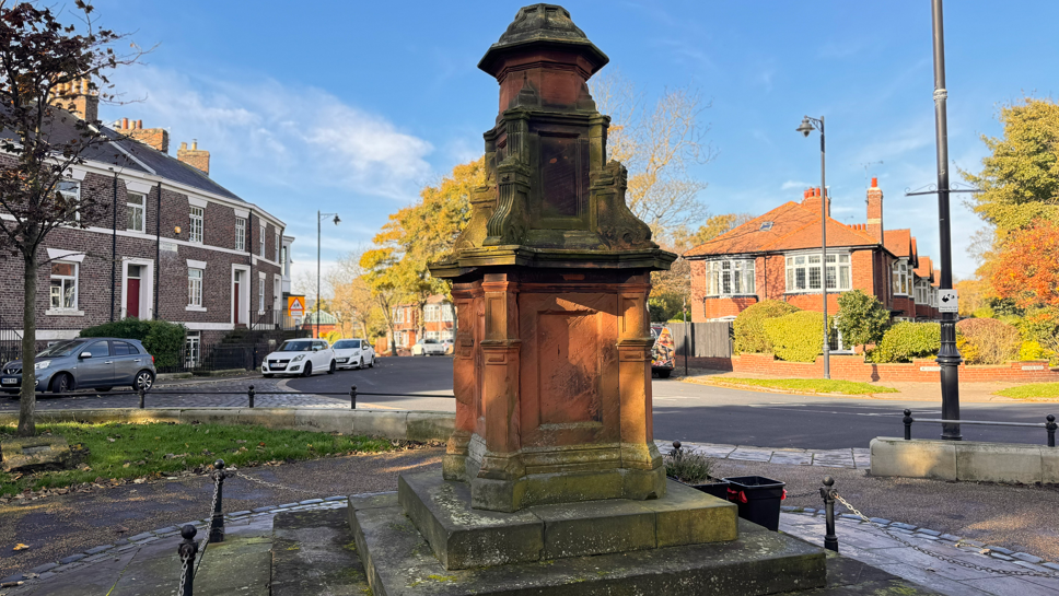 The Boer War memorial is orange and grey. A columns of descending sizes are stacked on top of each other on top of a plinth. It sits in a square surrounding by residential terraced houses.