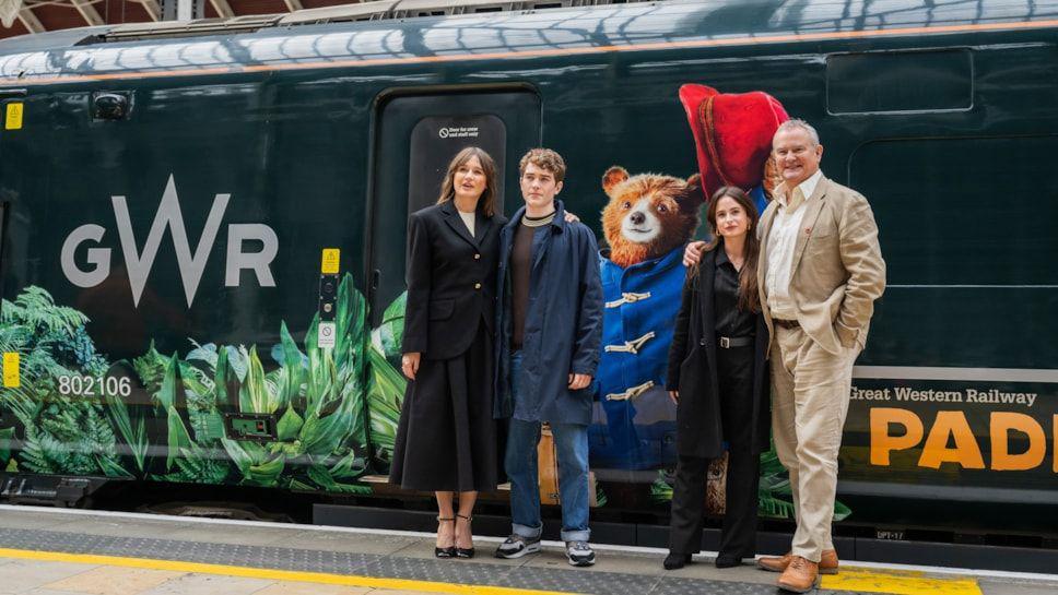 The stars Hugh Bonneville, Emily Mortimer, Madeleine Harris and Samuel Joslin pictured outside the train. The dark green train has tropical leaves painted alongside it with the words "Paddington" in orange capital letters.