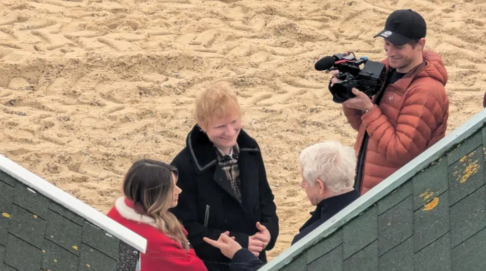 Ed Sheeran - a man with ginger hair wearing a black jacket - standing on a beach next to a woman wearing a red jacket and a man with a camera and a cap. They are talking to an elderly man with grey hair.
