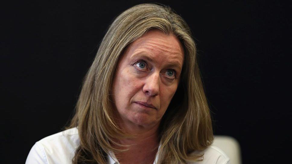 Justine Roberts, with brown shoulder-length hair and wearing a white top, looks ahead against a black background