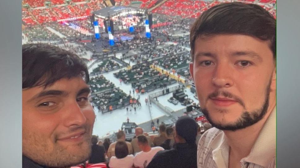 Ashwin and Connor take a selfie style image and smile. They are sat in the seats of a large arena. In the middle of it a boxing ring can be seen surrounded by seats.
