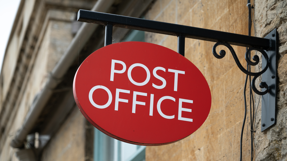 A post office sign is hanging off the side of a stone wall.