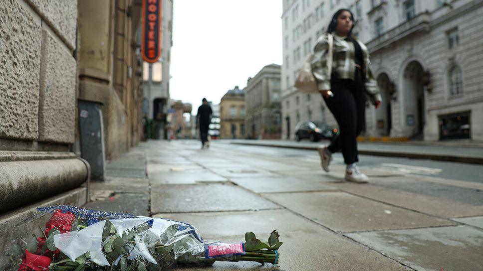 Red roses lie at Water Street by the site of an incident where a car ploughed into a crowd of Liverpool fans during a parade celebrating their side's Premier League win with a woman walking past.