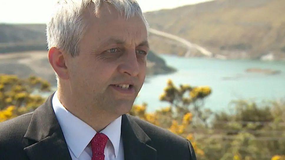 Dick Cole is talking in front of a lake and hillside. He has white hair and blue eyes and is wearing a black jacket, white shirt and red tie.