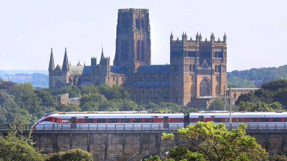 A grey and red LNER train passing on a viaduct on its way to Durham station. The line overlooks Durham Cathedral. It is a large stone building with several towers and intricate ornaments.