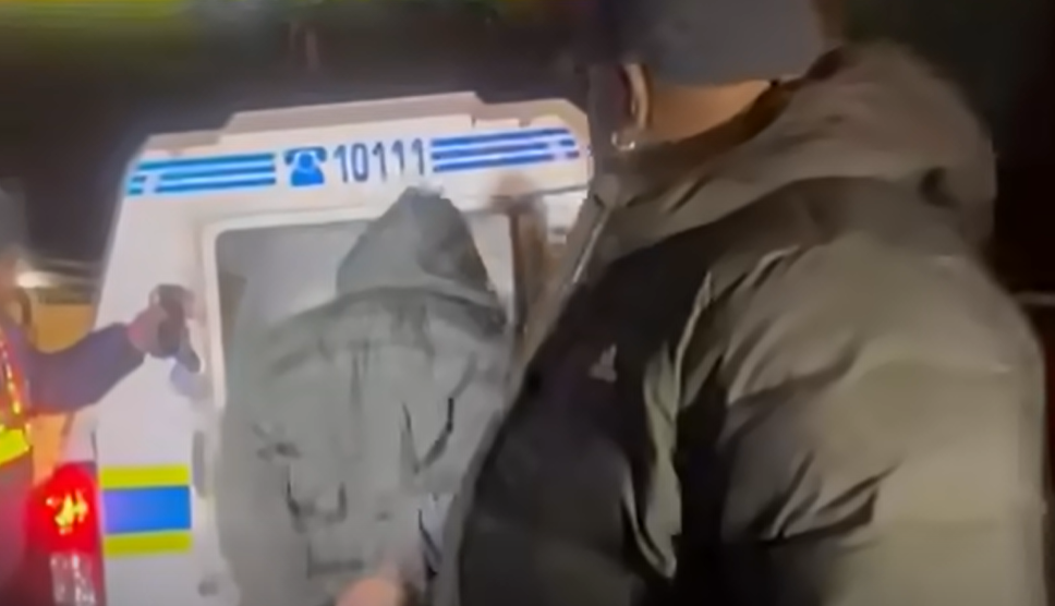 A man in a puffy coat and dark-coloured baseball cap is in the foreground, but looking away from the camera, towards someone in a grey coat being led into a police van. It is dark.