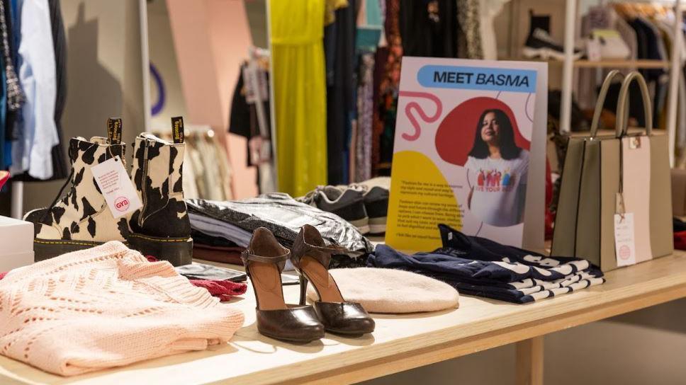 A close-up of one of the tables with donated items on it. It includes a pair of cow-print Doc Martens boots, brown heels, a pink jumper and light coloured beret. A multi-coloured sign is also visible telling the story of a client called Basra