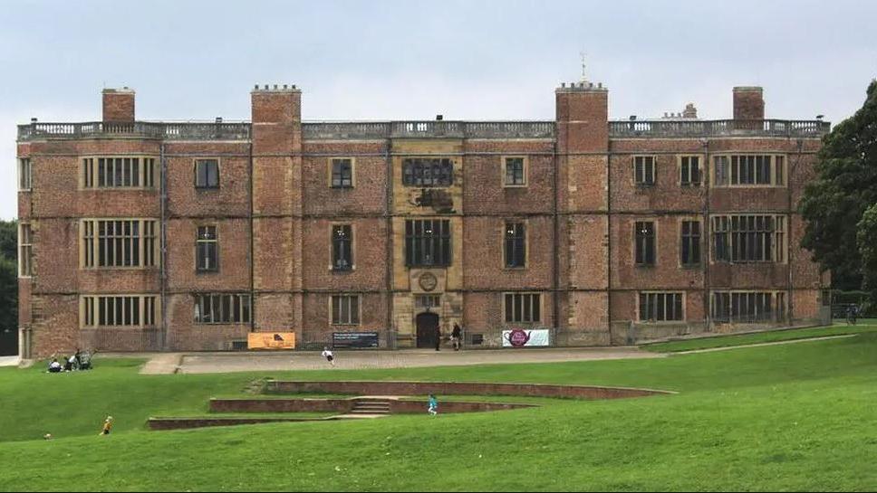 Temple Newsam, a Grade II listed Tudor-Jacobean house in Leeds. The building is red brick and has a large frontage and balustrade on the top. Gardens surround and children play outside.