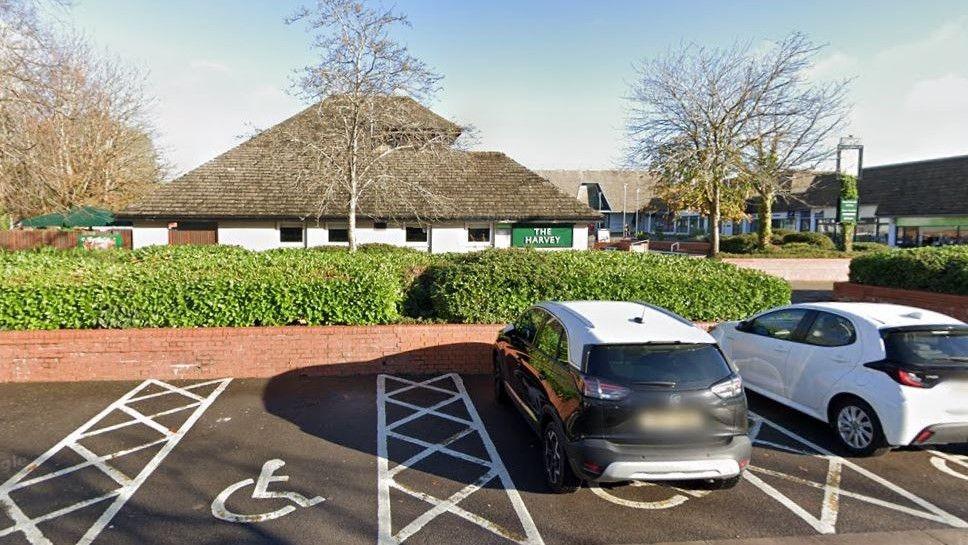 A Google Maps image of The Harvey Pub, taken from the car park. There is a green sign on the building which has trees in front of it.