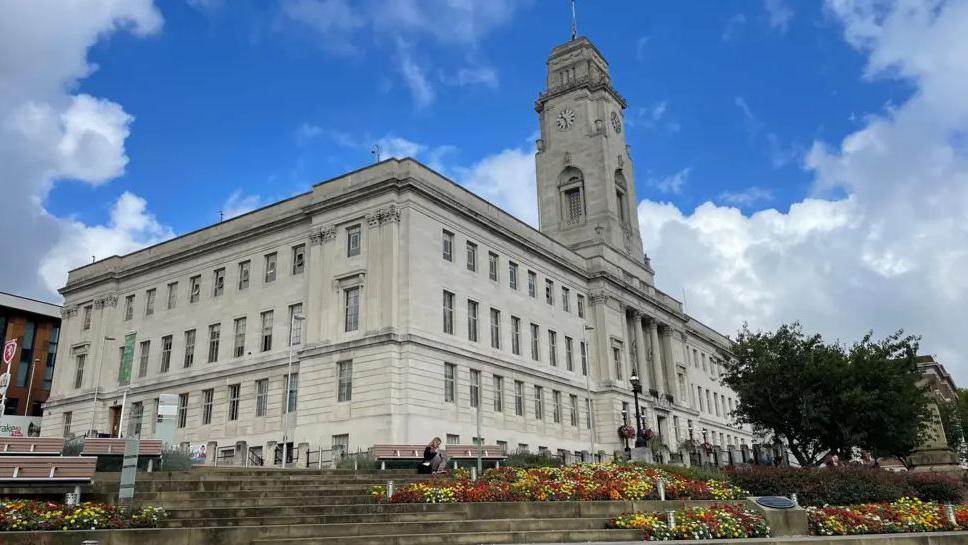 A large council building with four storeys and a clock tower, with flower beds and steps at the front. 