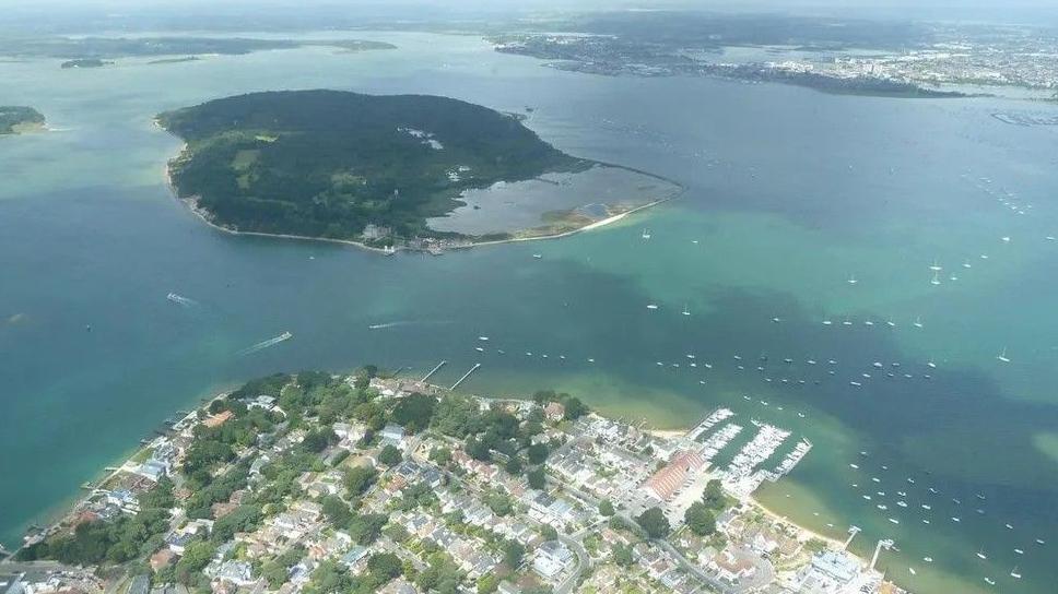 Poole Harbour from the air, with houses on the coast at the bottom of the picture. The heavily-forested Brownsea Island is at the top.