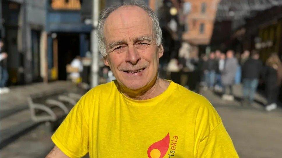 A man, Séamas Ó Labhradha pictured outside in Belfast city centre. He has grey hair and is wearing a yellow top with the arts organisation's logo Arstekta on it in red and black.