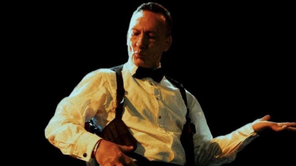 Piers Burnell dressed in a suit like James Bond poses for an air guitar performance. His eyes are closed.