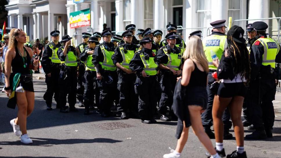 A group of police officers in hi vis and police hats patrol on the streets of Notting Hill as revellers walk past