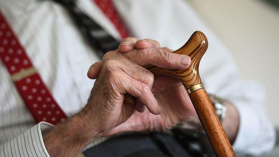 The hands of an elderly man, dressed smartly in a white striped shirt, dark tie and red braces and holding the handle of a wooden walking stick.