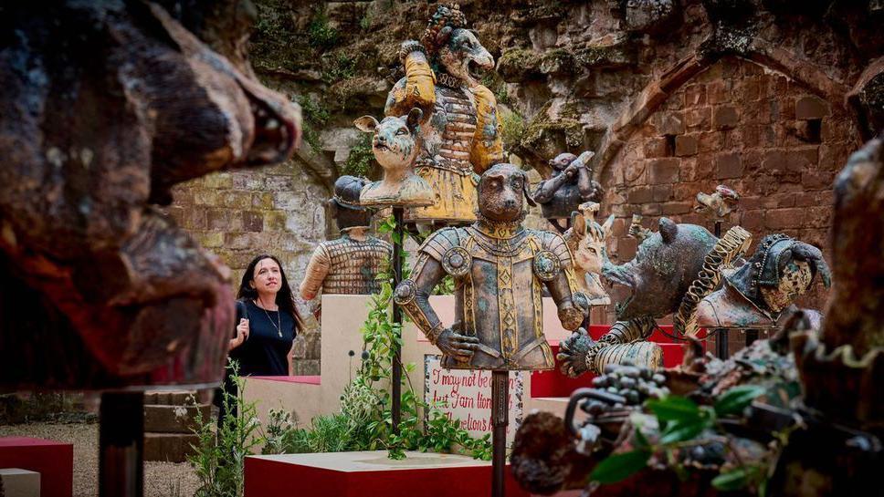 A woman with dark hair and a dark top in a large outdoor area, surrounded by a collection of unusual decorative statues and busts 
