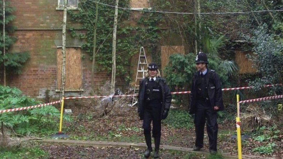 A female and male police officer in uniform are pictured in the grounds of a house. There are bushes and trees in the background and some windows on a building are boarded up.