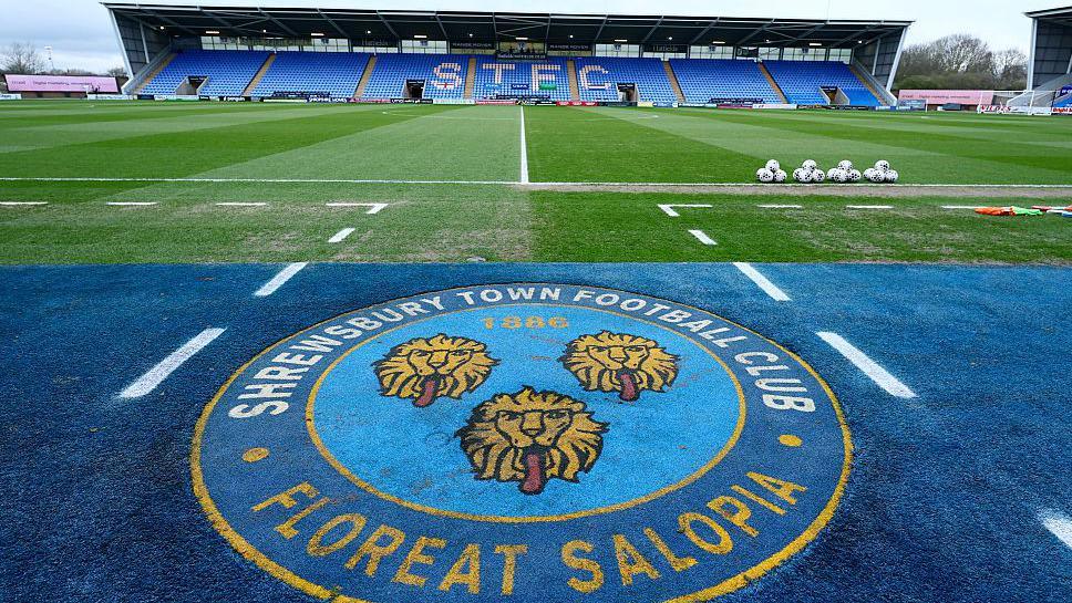 The Shrewsbury Town club crest with a stand at the Croud Meadow in the background