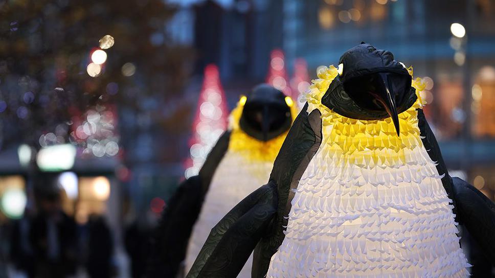 Giant penguin characters with Liverpool ONE shopping centre blurred out in the background.