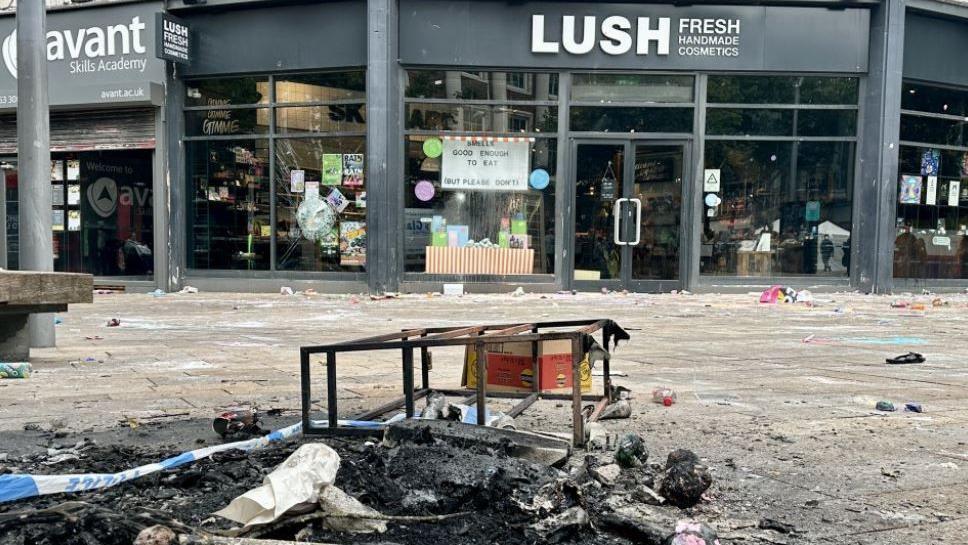 Several burnt items on the street and a metal railing with a police cordon sign on the floor and Lush in the background