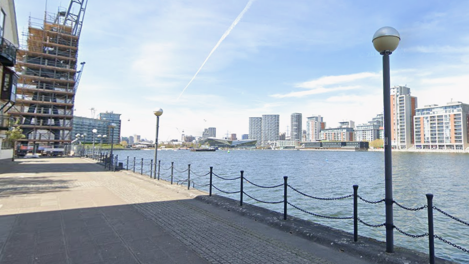 Waterfront walkway beside Royal Victoria Dock, with railings, lamp posts, modern flats and a building under construction on a clear day.