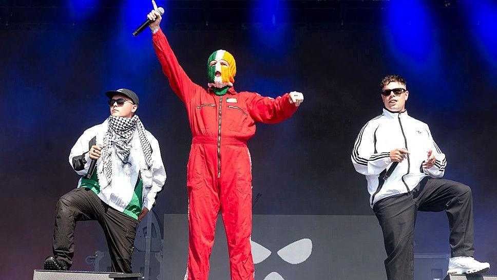 Three members of Belfast rap trio Kneecap on stage. Mo Chara, DJ Provai and Moglai Bap all carry microphones. Mo Chara wears a black baseball cap, black sunglasses and a white and green sports top and black sports trousers and black trainers. He has a traditional Palestinian keffiyeh shawl wrapped around his shoulders. DJ Provai wears a red boilersuit and has a balaclava in green, white and orange over his head. He has his hand with the microphone raised over his head. Moglai Bap wears black cap and sunglasses, a white Adidas long-sleeved jacket, black sport trousers and white trainers.