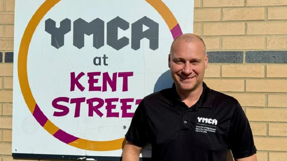 Rob Baty stood in front of the "YMCA at Kent Street" sign, smiling at the camera and wearing a black T-shirt with "YMCA" written on in white letters.
